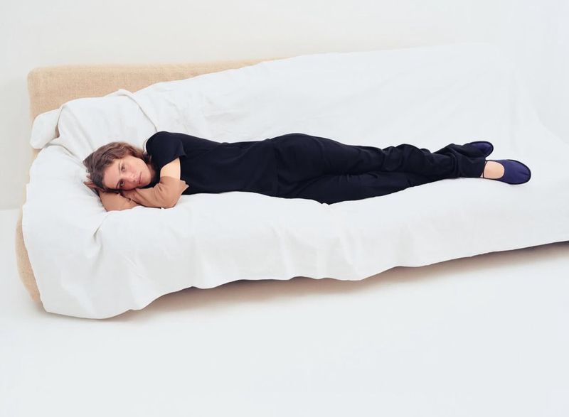 An adult is lying on a white couch with a tan-colored back cushion in a brightly lit indoor space, wearing a dark outfit and blue slippers.