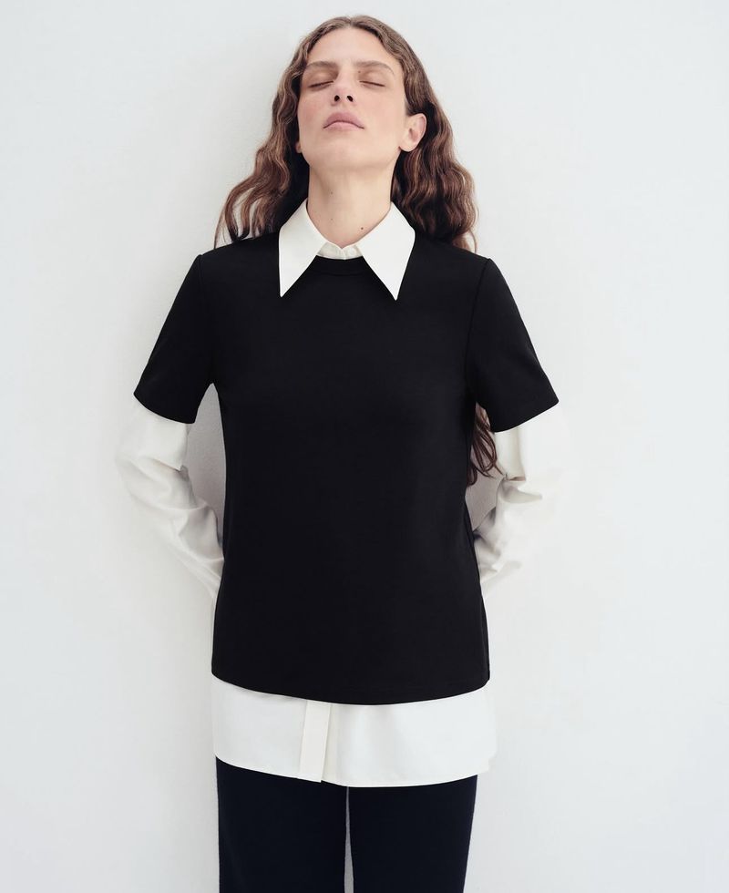 Fashion shot of a woman in a black top with a white collar and long sleeves, posing against a plain white background in a studio.