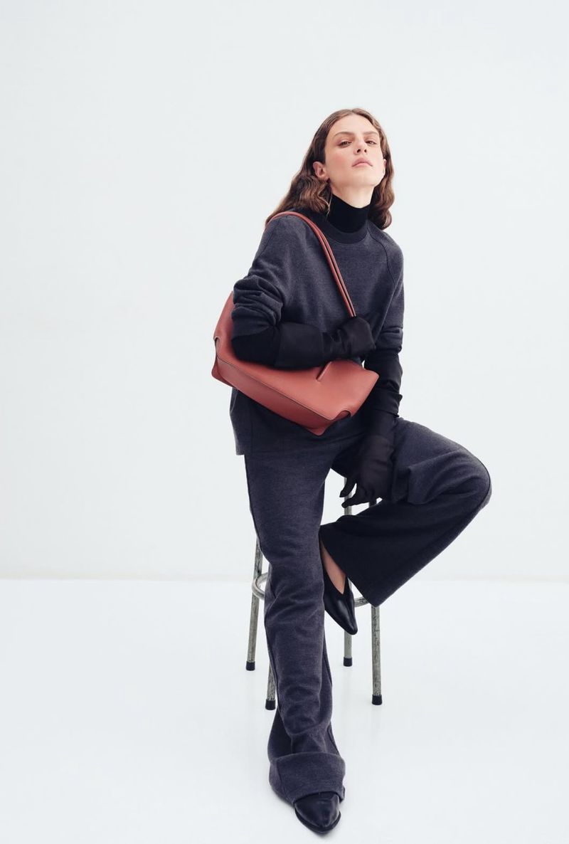 A fashionable woman poses indoors with an orange purse and a gray outfit, seated on a stool against a bright white studio backdrop.