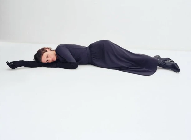 Model lying horizontally on a white floor wearing a dark long-sleeve top, matching skirt, black opera gloves, and boots.