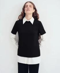 Model with eyes closed in a layered black short-sleeved top over a white collared shirt against a white backdrop.