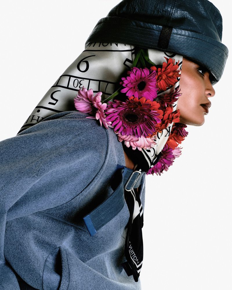 Fashionable woman in blue hat and jacket adorned with flowers and scarf, striking pose against a clean white background.