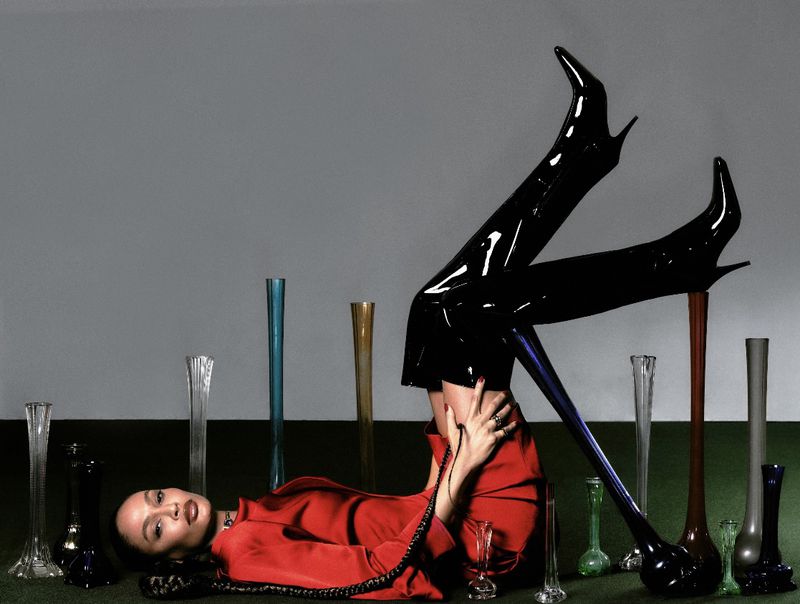 A woman lies on the floor in a red top, black patent leather boots, and jewelry surrounded by tall vases against a grey wall.
