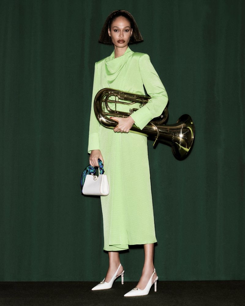Fashion model in a light-green dress holds a tuba and white purse against a green curtain backdrop, accessorized with white heels.