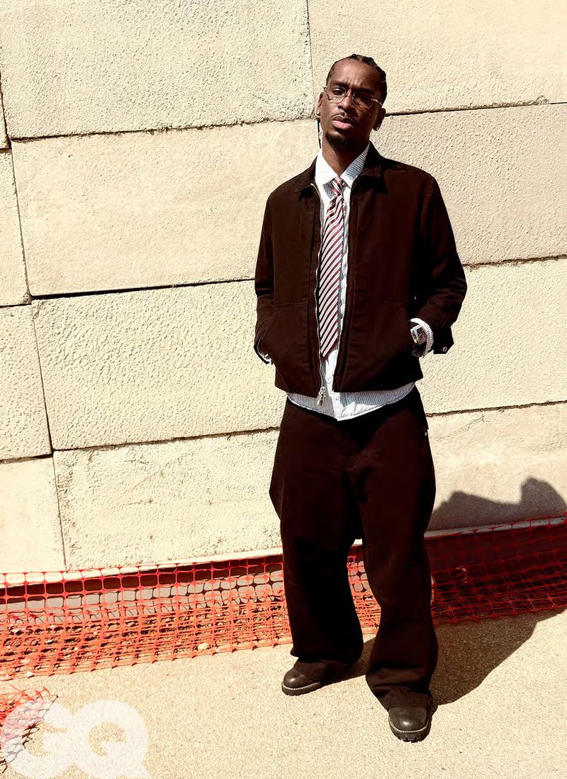 Adult male with glasses poses outdoors near a concrete wall in brown jacket, pants, dress shirt and tie for a fashion editorial shot.