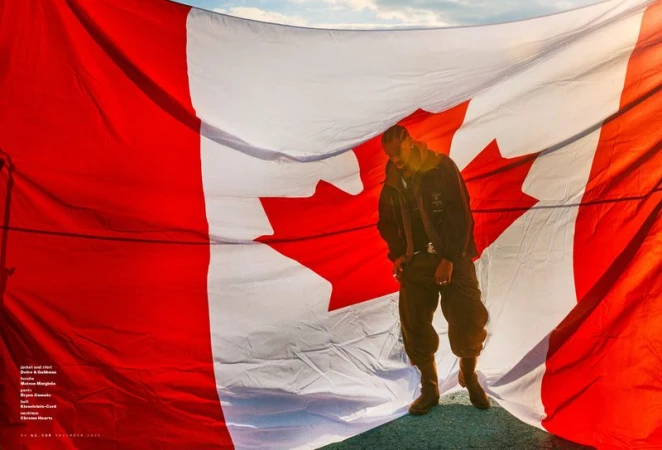 Man posing in front of a large Canadian flag wearing a jacket and pants, set against a bright outdoor setting with a blue sky.