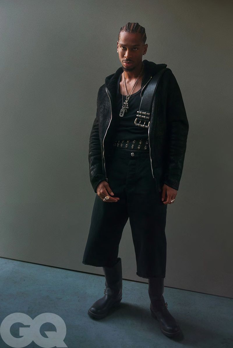 Portrait of a Black man in black culottes and a black hooded jacket in a studio, wearing jewelry, against a grey backdrop.