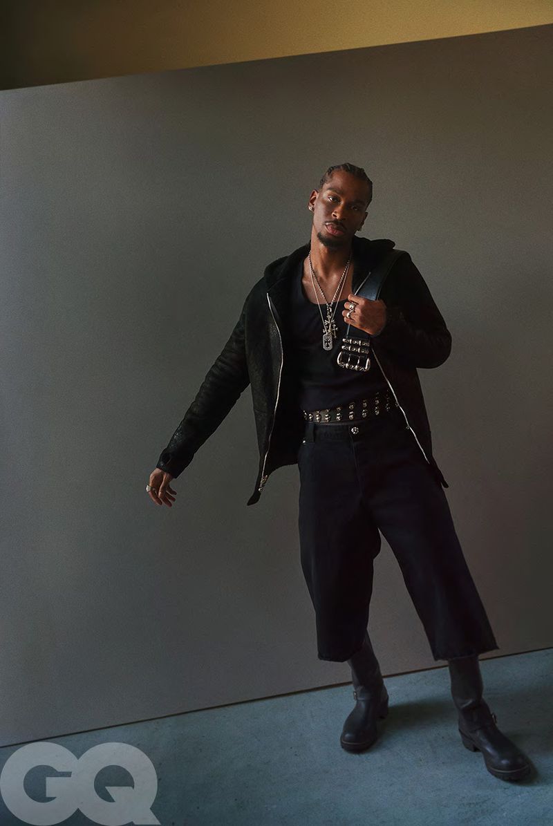 A full portrait of a man in a black outfit. He wears a jacket, pants, and boots, accessorized with jewelry. The background is neutral grey.