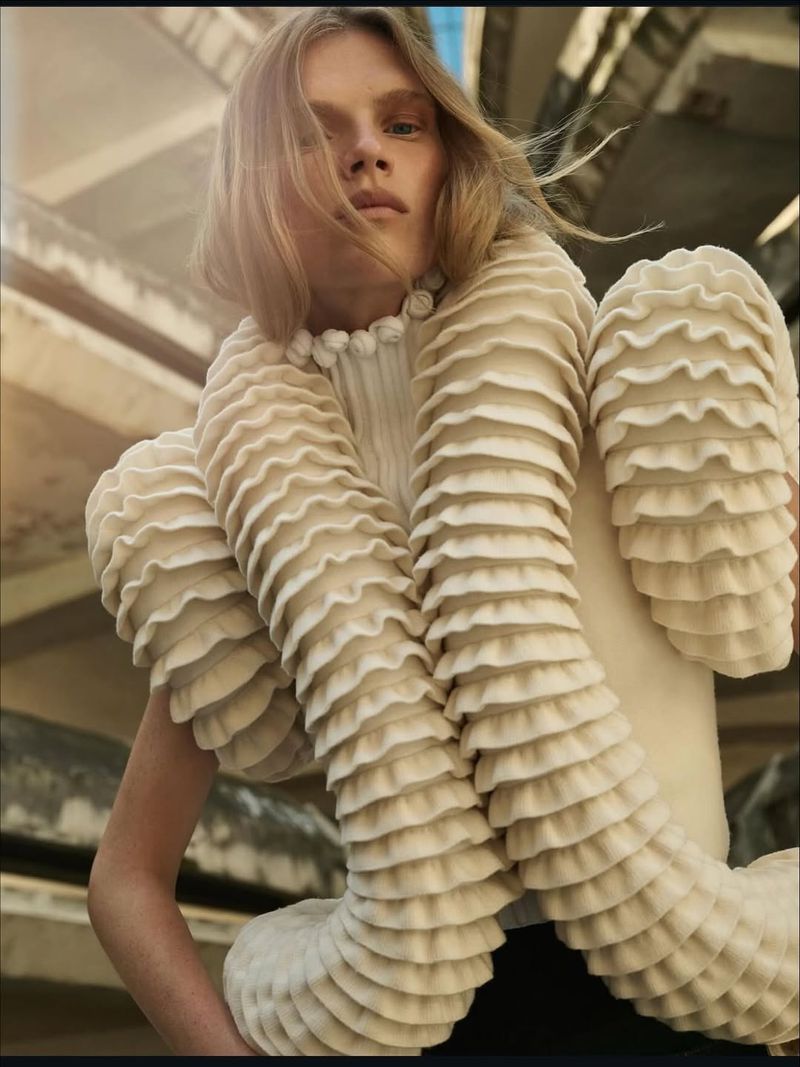 Woman in a ruffled cream-colored top poses in a low-angle shot with a weathered urban structure providing depth to the background.