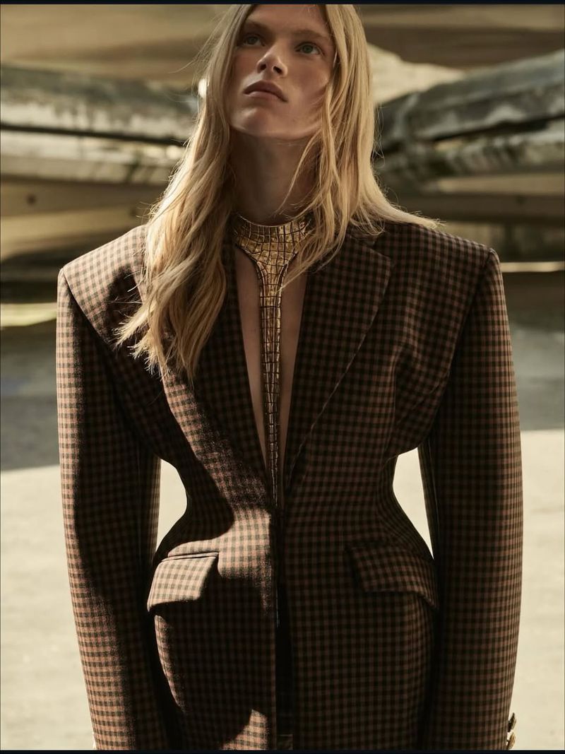 Fashion shot of a woman with long blonde hair, wearing a patterned blazer and gold necklace in a studio with a rocky background.