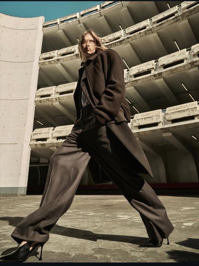 Fashion shot of a blonde woman in a brown coat and trousers walking on a concrete surface in front of a concrete building.