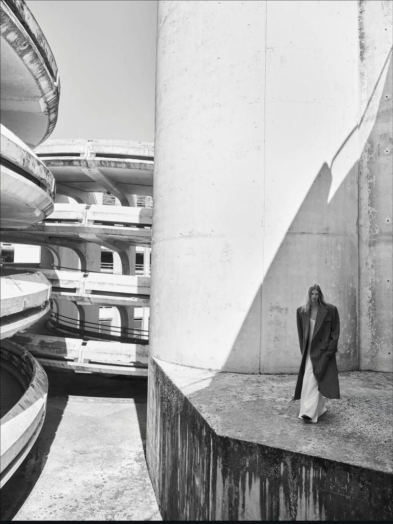 Monochrome portrait of a woman in a long coat, standing in front of a concrete wall, with architectural structure in the background.