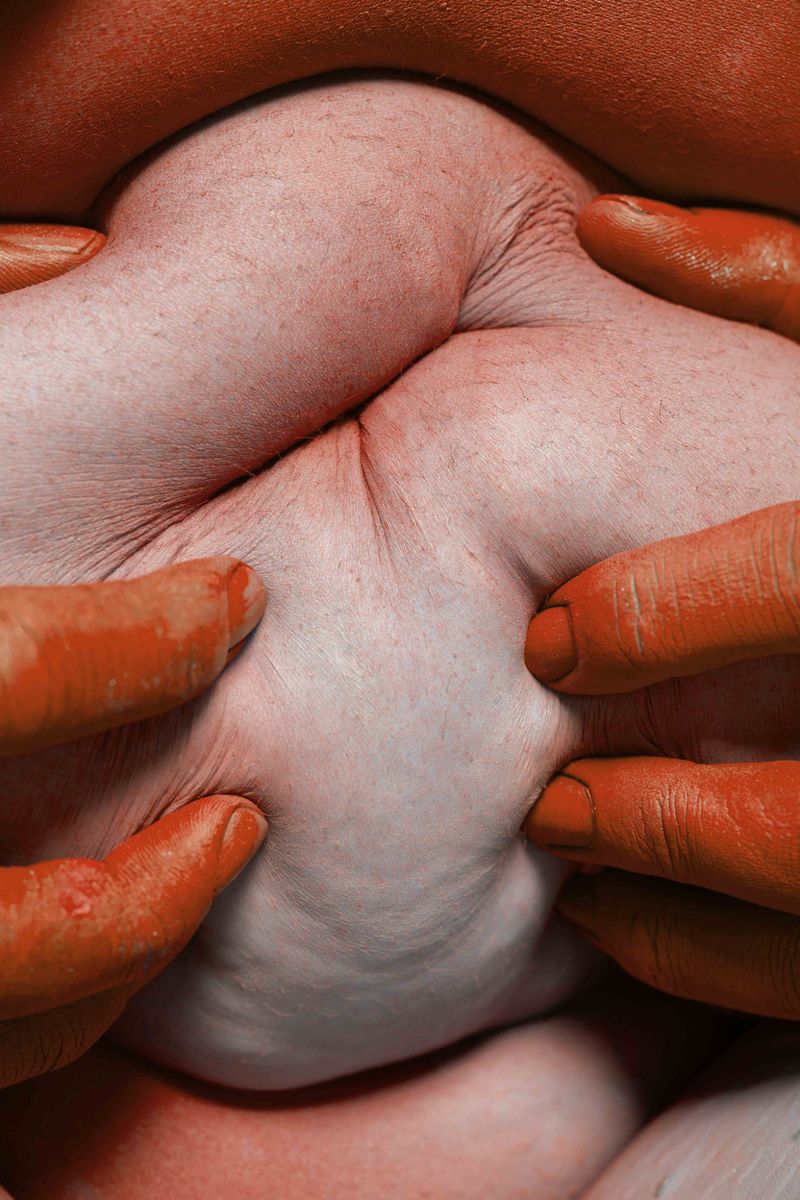 Close-up shot of a baby being held by two adults, displaying fingers and arms against a muted orange-toned background.