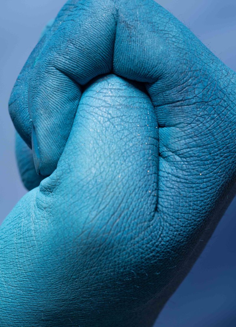 Close-up of a fist covered in teal-blue paint against a blurry, graded blue background, showing skin texture and paint consistency.