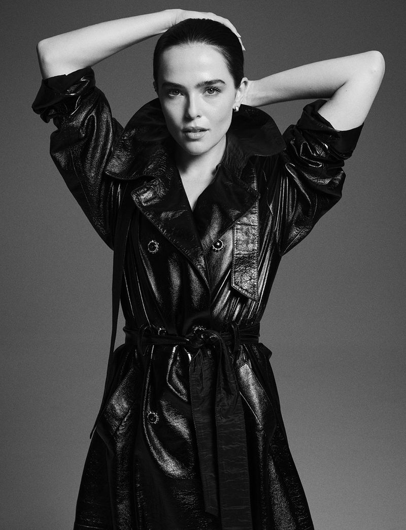 Monochrome portrait of a woman with hands behind her head, wearing a shiny black trench coat against a plain gray background.