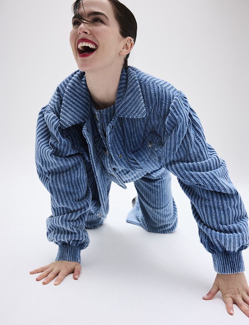 A young woman with dark hair, smiling widely while kneeling and leaning forward, wearing a blue corduroy jacket and pants on a white backdrop.