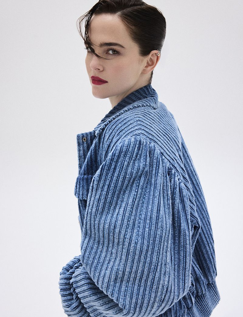 Portrait of an adult female with red lipstick, wearing a light blue jacket with large sleeves, against a white background, looking back.