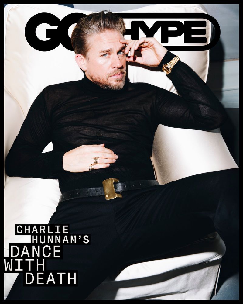 Charlie Hunnam on a magazine cover, wearing a black turtleneck and suit, gold watch, and rings, posed on a cream colored armchair.
