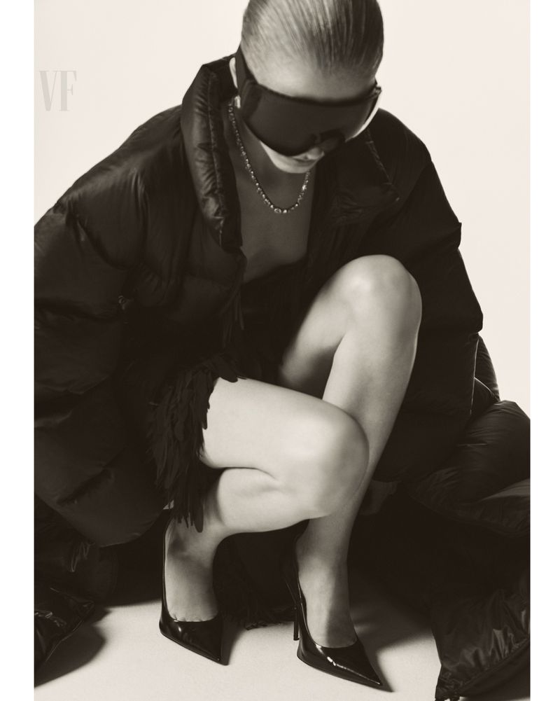 Woman in puffy coat and sunglasses kneeling in studio wearing heels. Fashion-style studio shot with neutral color palette, soft lighting.