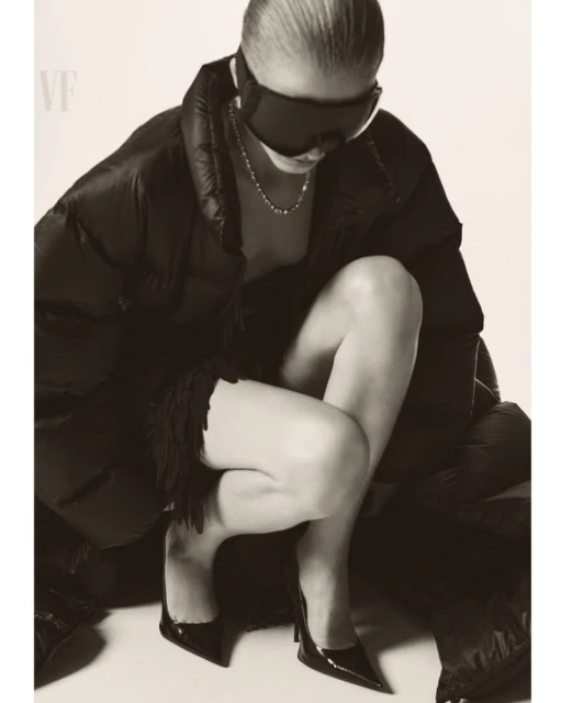 Woman in puffy coat and sunglasses kneeling in studio wearing heels. Fashion-style studio shot with neutral color palette, soft lighting.