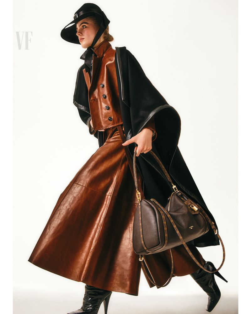 Fashionable woman in a brown leather skirt, vest, and hat, paired with a black cloak and boots, carrying a brown leather handbag.