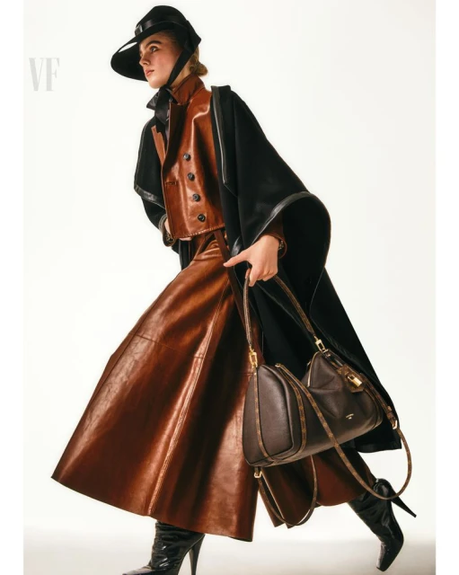 Fashionable woman in a brown leather skirt, vest, and hat, paired with a black cloak and boots, carrying a brown leather handbag.