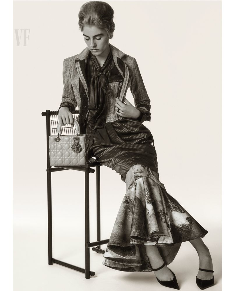 Monochrome fashion shot of a model in a tailored jacket and long skirt, sitting on a black chair with a purse on the chair.