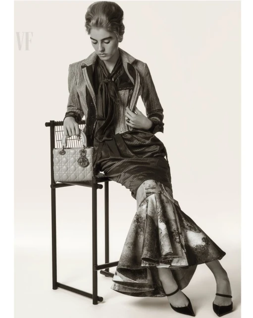 Monochrome fashion shot of a model in a tailored jacket and long skirt, sitting on a black chair with a purse on the chair.