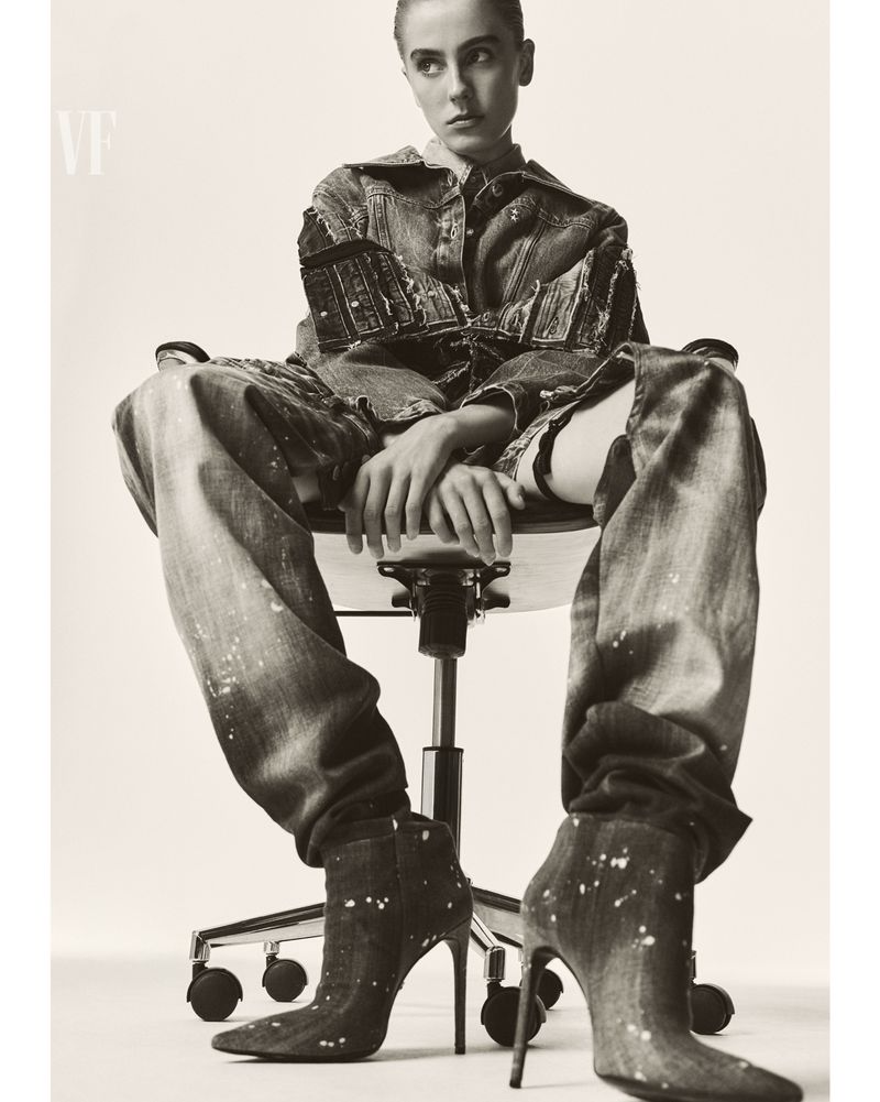 Fashionable young adult woman poses in a light studio wearing a denim jacket, jeans and boots with high heels, sitting in an office chair.