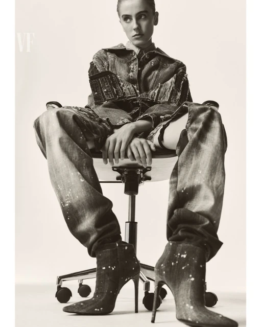 Fashionable young adult woman poses in a light studio wearing a denim jacket, jeans and boots with high heels, sitting in an office chair.