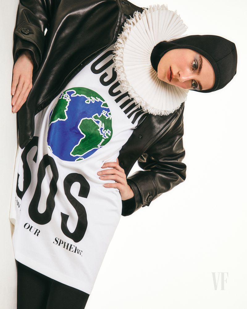 A person wearing a Moschino t-shirt with an Earth graphic and the letters 'SOS' in black, and a black leather jacket, against a white wall.