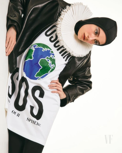 A person wearing a Moschino t-shirt with an Earth graphic and the letters 'SOS' in black, and a black leather jacket, against a white wall.