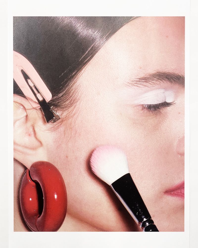 Close-up of a woman's face with white eye makeup, red earrings, and a makeup brush applying blush, set against a white background.