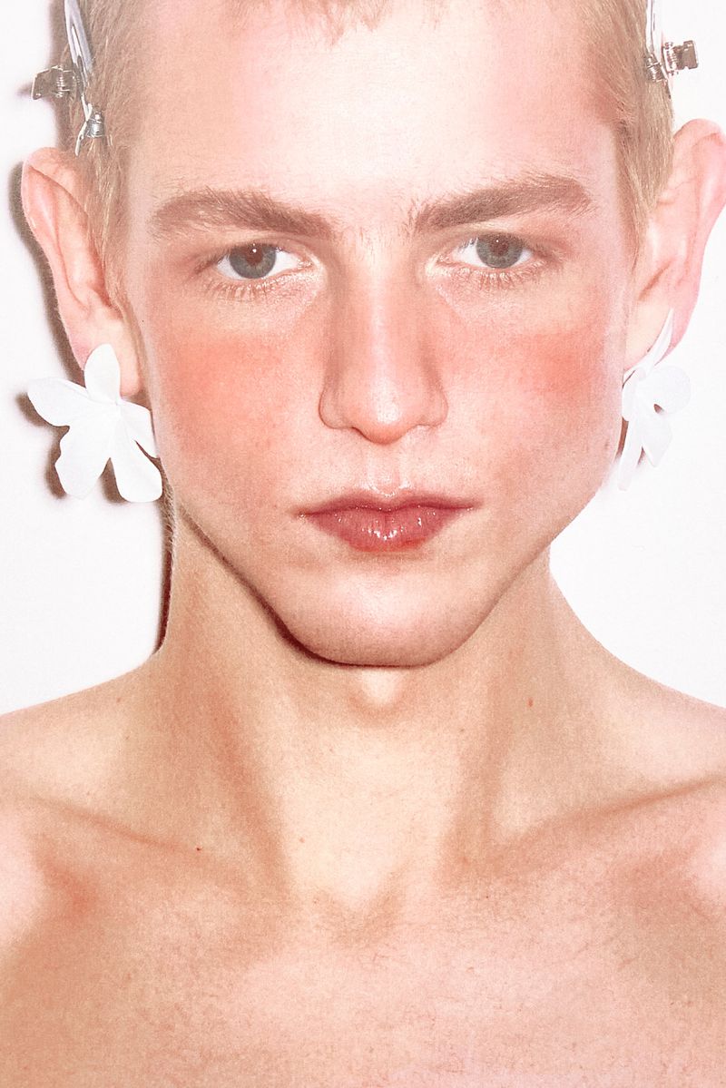 Close-up portrait of a young adult with short blond hair, wearing large flower earrings against a white background in a beauty shot.