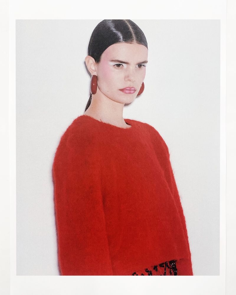 A woman poses in a red sweater and dark skirt with large earrings, standing in front of a white wall in a studio portrait.