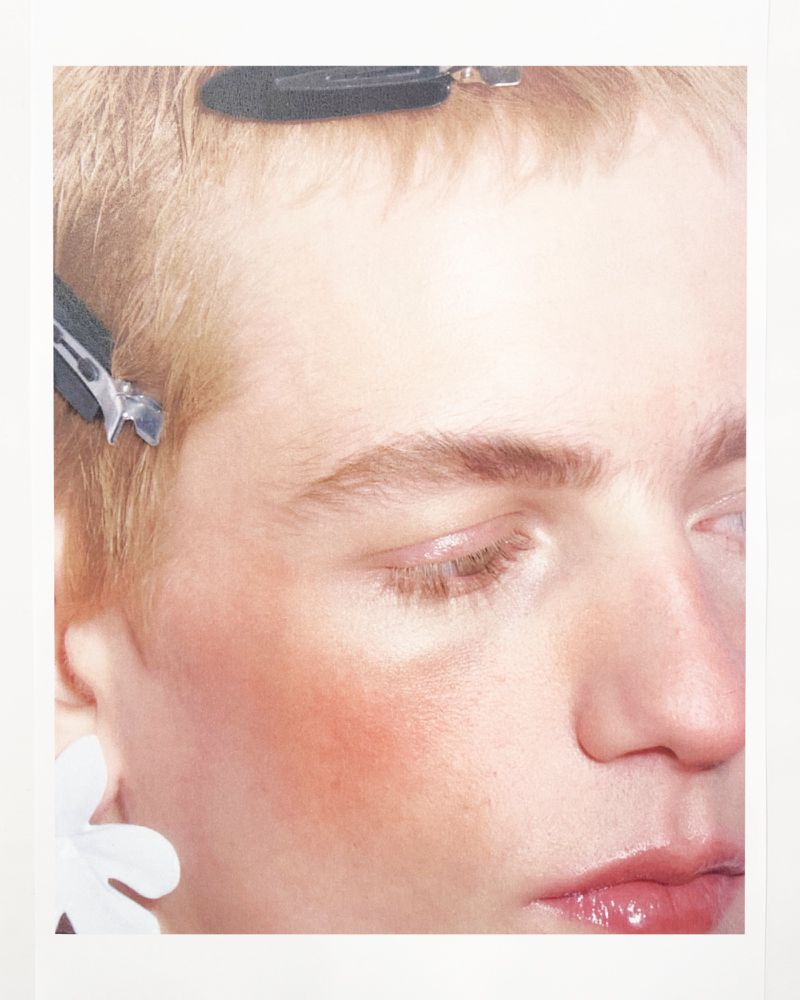 Close-up portrait of a person with fair skin, light hair, and hair clips, featuring soft lighting and a minimal background.