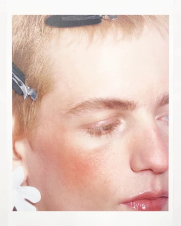 Close-up portrait of a person with fair skin, light hair, and hair clips, featuring soft lighting and a minimal background.