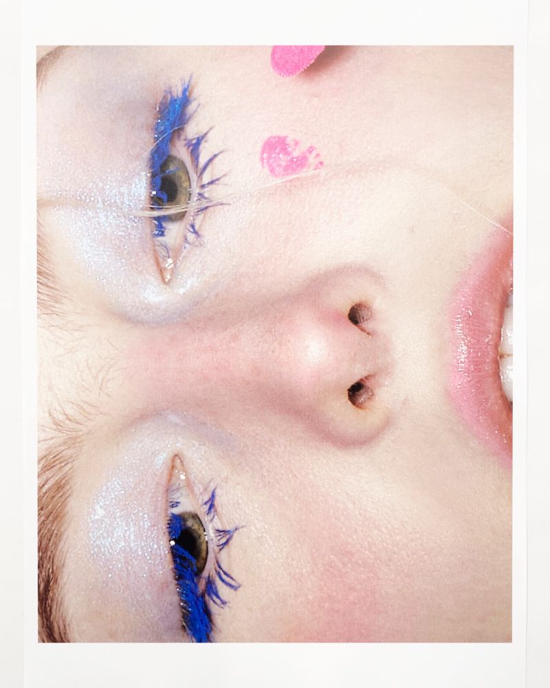 Close-up portrait of a fair-skinned woman with blue mascara on her lower lashes and pink heart-shaped decorations on her face.