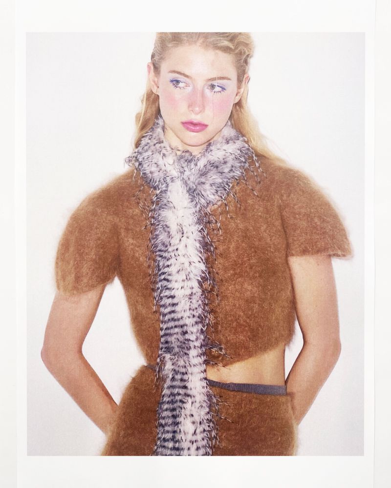 Portrait of a blonde woman with light makeup wearing a brown knit outfit with a feathered scarf against a white background.