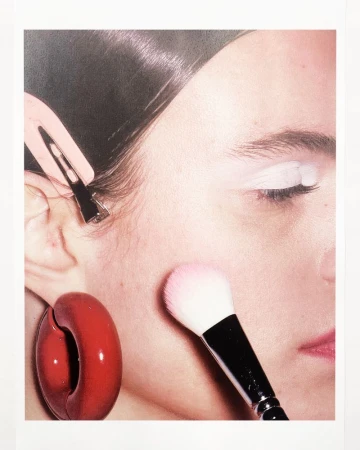 Close-up portrait of a model having blush applied with a makeup brush, featuring a pink hair clip and red hoop earring.