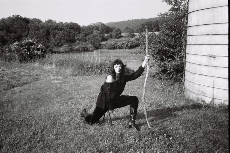 A woman in a black outfit poses with a walking stick in a grassy field near a cylindrical silo, creating a monochromatic outdoor scene.