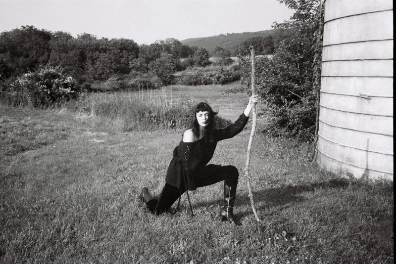 A woman in a black outfit poses with a walking stick in a grassy field near a cylindrical silo, creating a monochromatic outdoor scene.