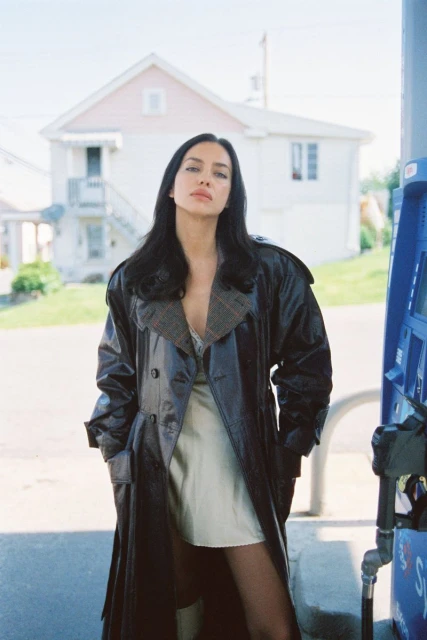 A woman in a dark trench coat poses in front of a vintage gas station and a white house on a sunny day in an outdoor setting.