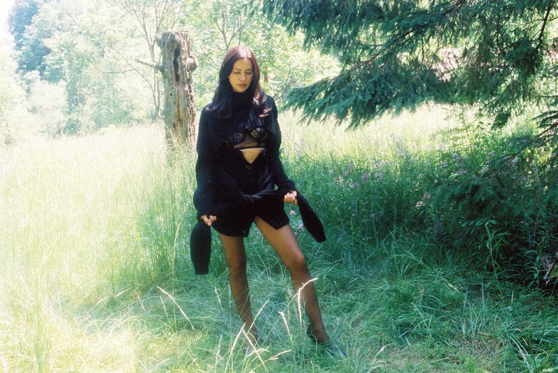 Young woman in black sweater stands in a sunlit grassy field next to a forest with green trees.