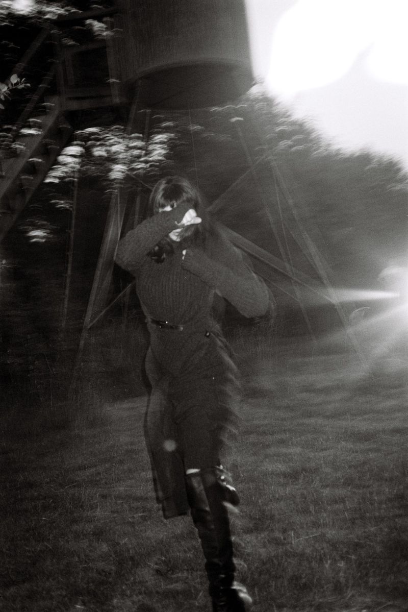 Monochrome photo of a woman in dark clothing running through a grassy field toward a water tower, with a motion blur effect.