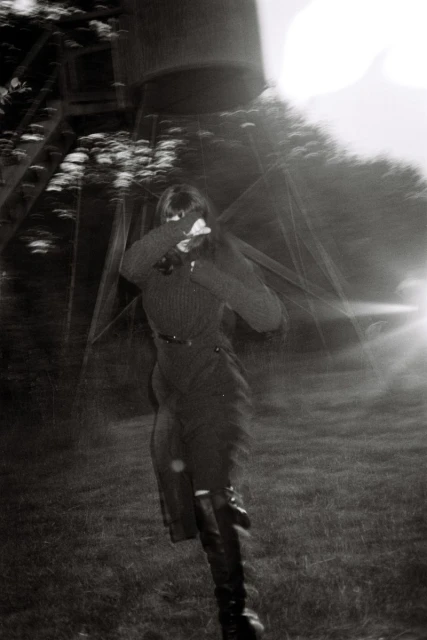 Monochrome photo of a woman in dark clothing running through a grassy field toward a water tower, with a motion blur effect.