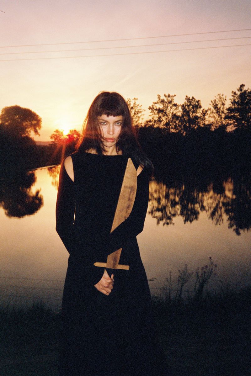 A woman in a black dress stands near a lake at sunset, holding a wooden sword with a determined expression and dramatic lighting.
