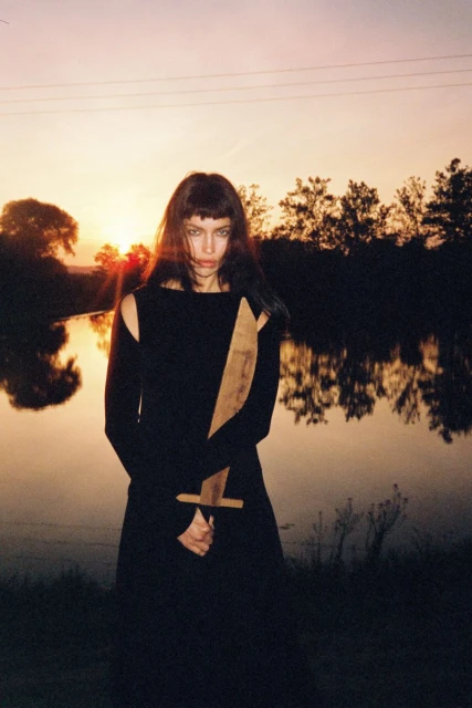 A woman in a black dress stands near a lake at sunset, holding a wooden sword with a determined expression and dramatic lighting.