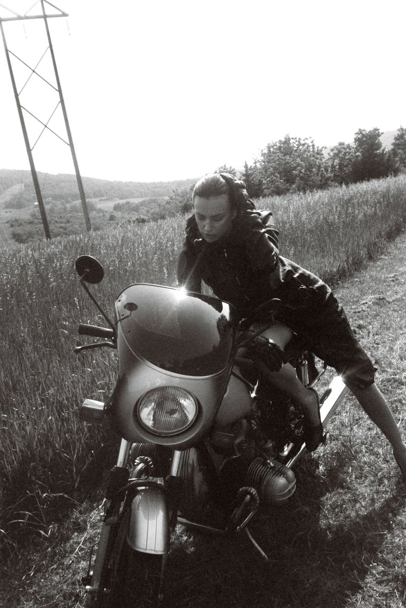 Monochrome shot of a woman in casualwear posing on a vintage motorcycle on a grassy path with power lines in the background.