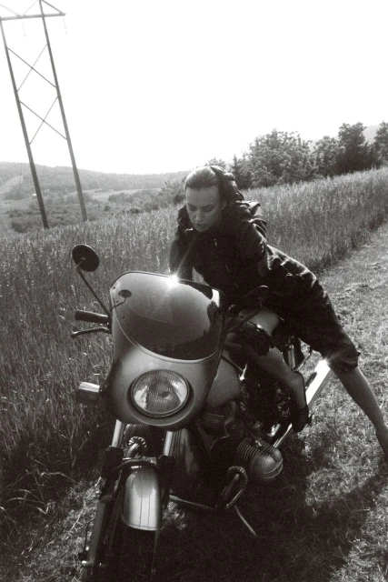 Monochrome shot of a woman in casualwear posing on a vintage motorcycle on a grassy path with power lines in the background.